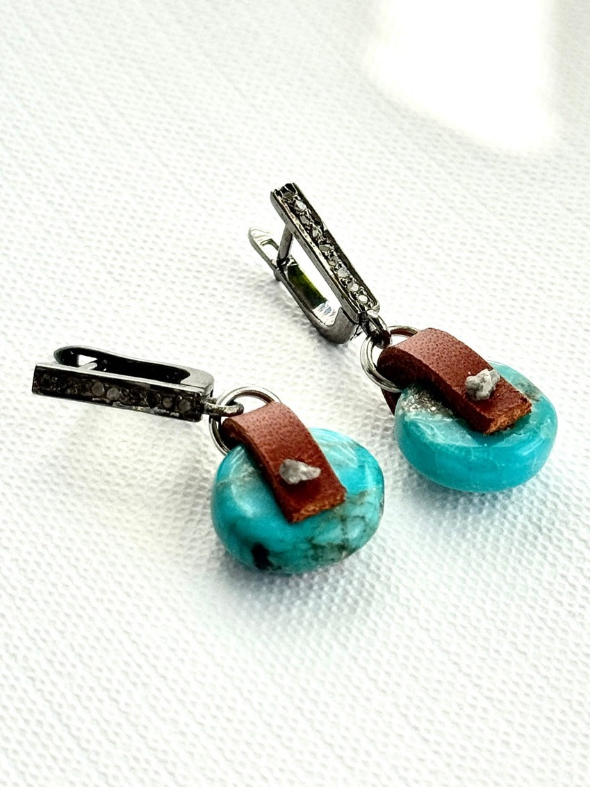 Arizona turquoise coin earrings with hand-sewn raw grey diamonds on chestnut brown leather, angled view lying flat