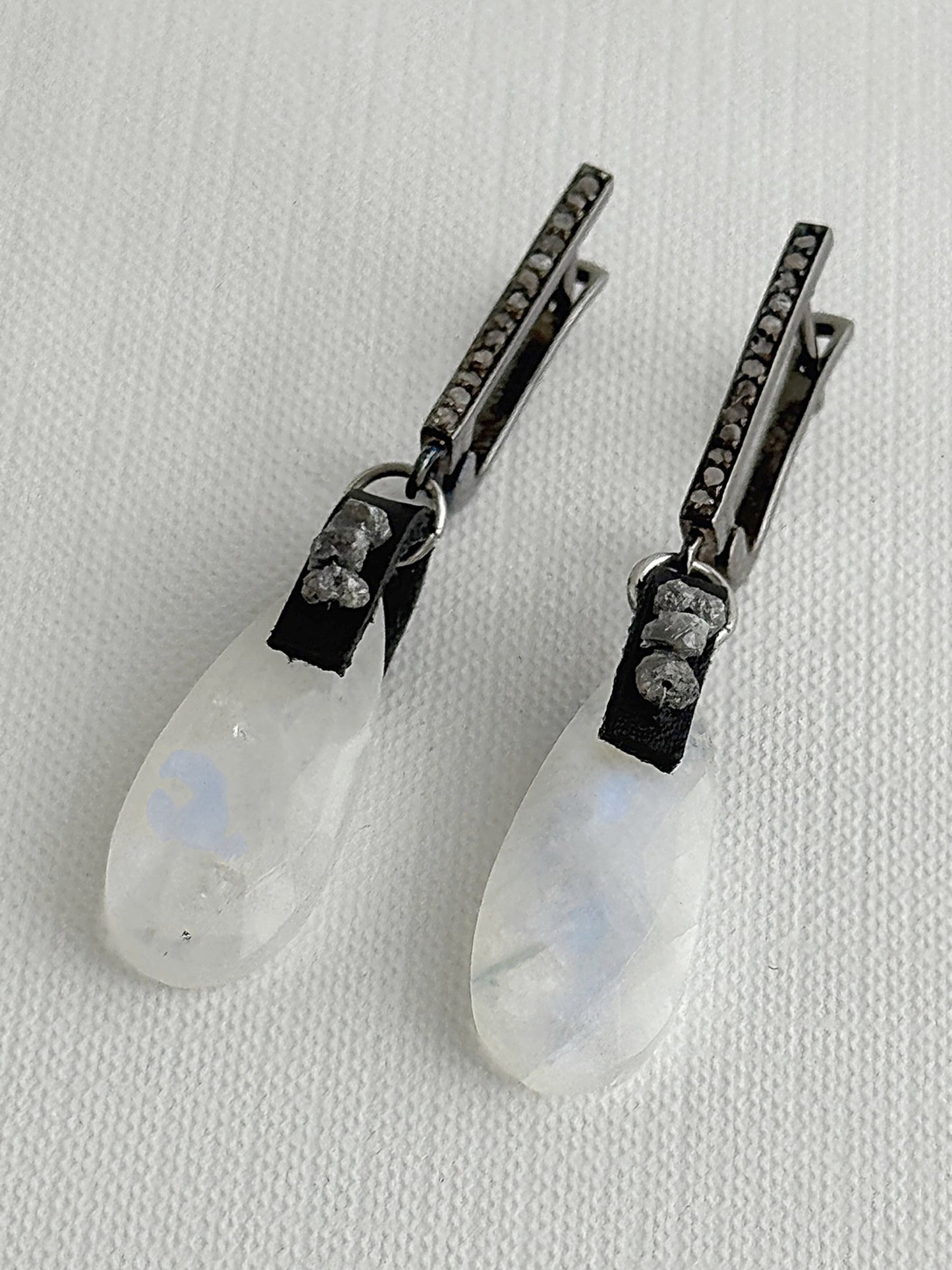 Angled view of rainbow moonstone teardrop earrings showing the blue flash, with raw grey diamonds on black leather and oxidized sterling silver pavé leverbacks