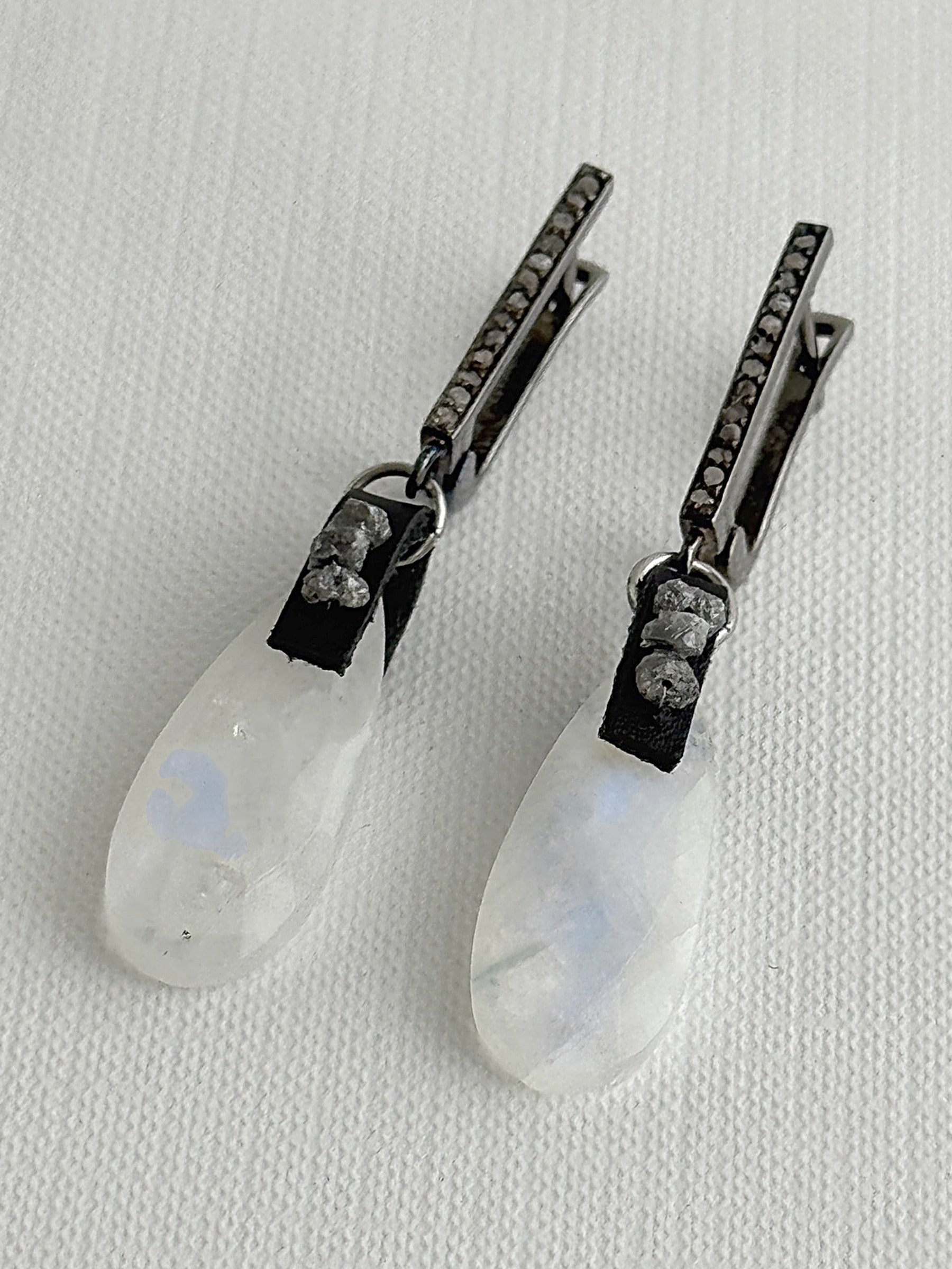 Angled view of rainbow moonstone teardrop earrings showing the blue flash, with raw grey diamonds on black leather and oxidized sterling silver pavé leverbacks