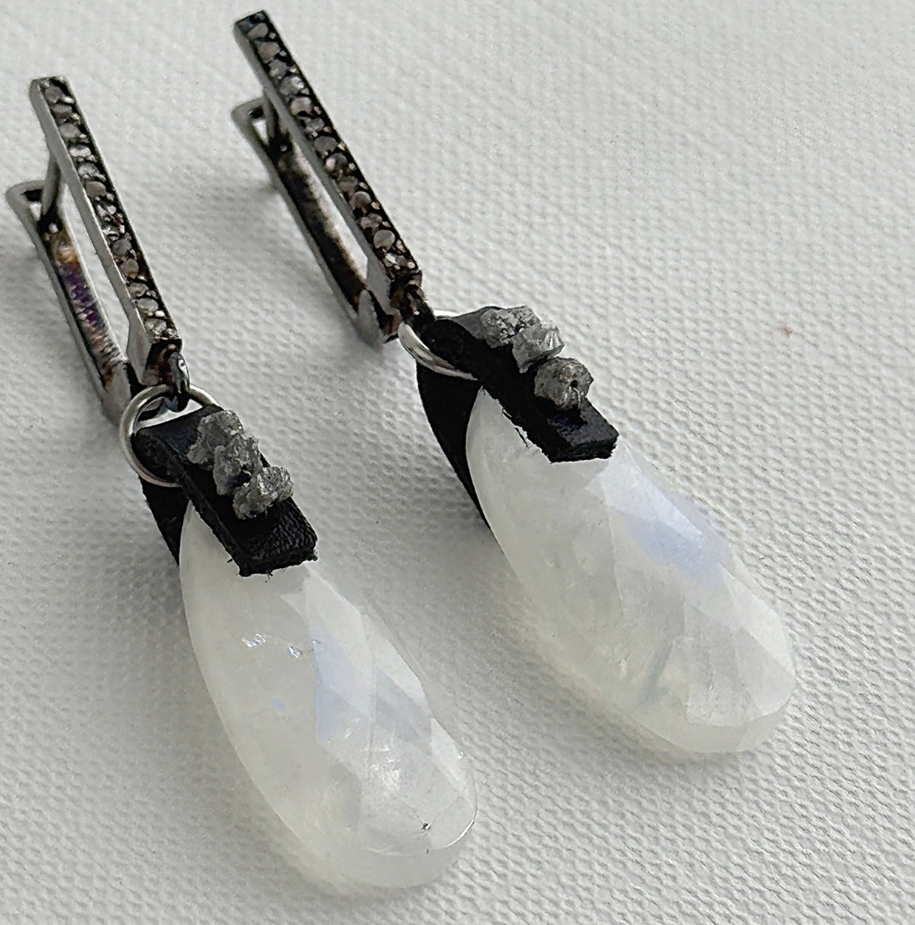 Close-up of rainbow moonstone teardrop earrings with raw grey diamonds on black leather and oxidized sterling silver pavé leverbacks