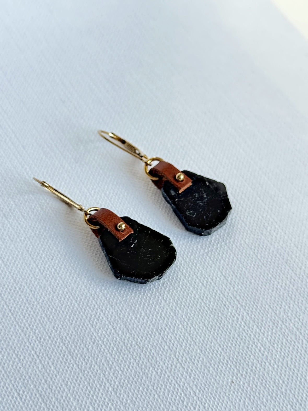 Glossy black tourmaline freeform slice earrings with a hand-sewn 14K gold-filled bead on brown leather and 14K gold-filled leverbacks, angled view